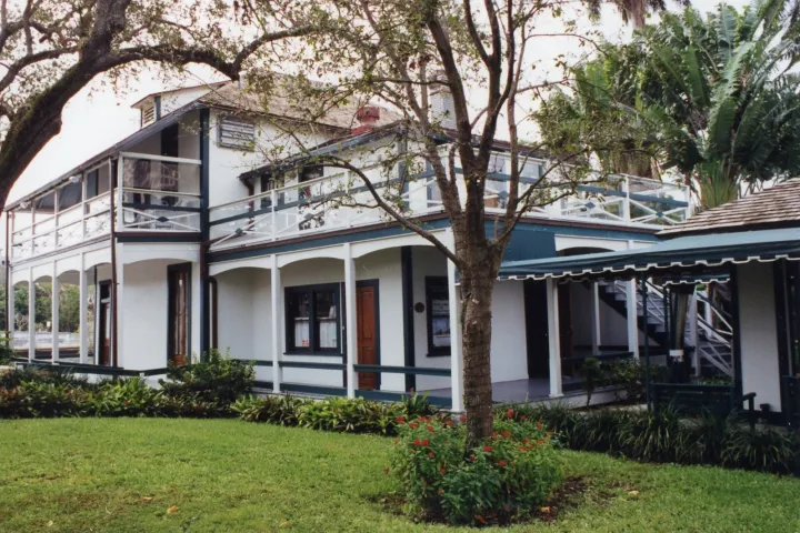 Two-story white building with a veranda and greenery in the foreground.