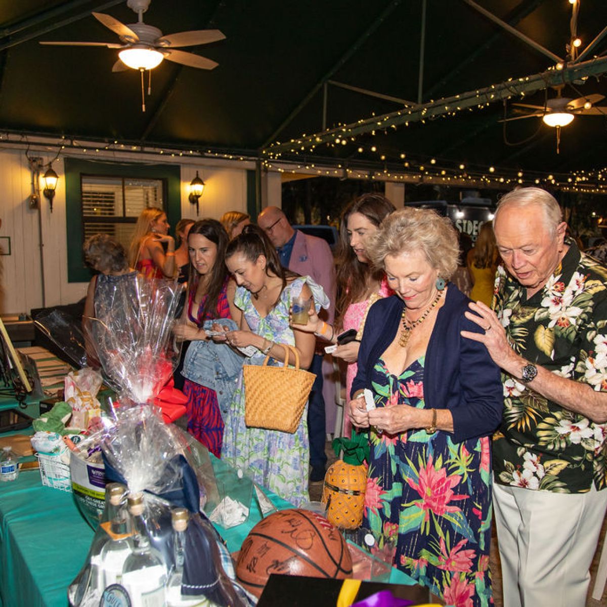 People gathered at a dimly-lit event, looking at a table with raffle items and auction baskets.