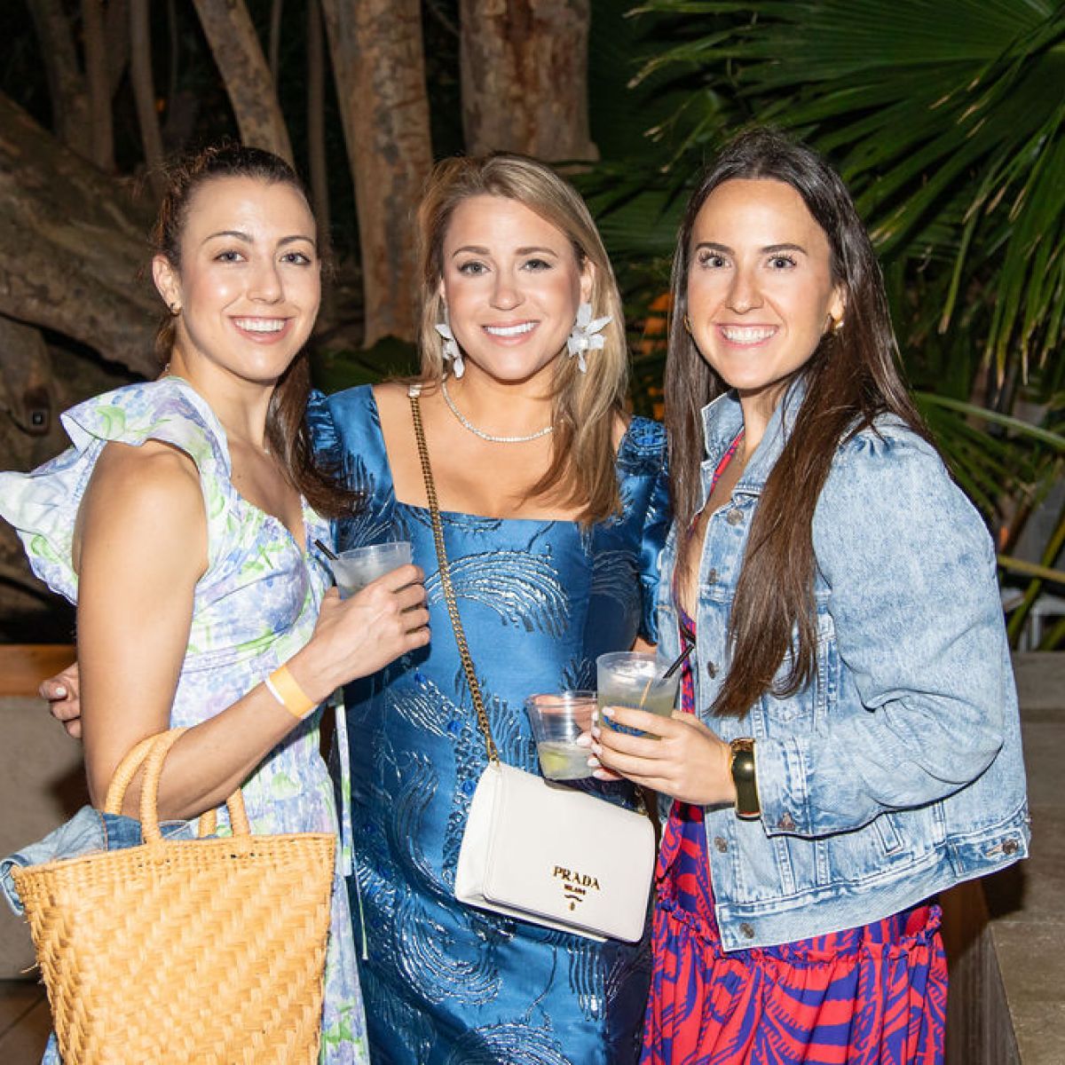 Three women smiling and holding drinks, standing outdoors near trees.