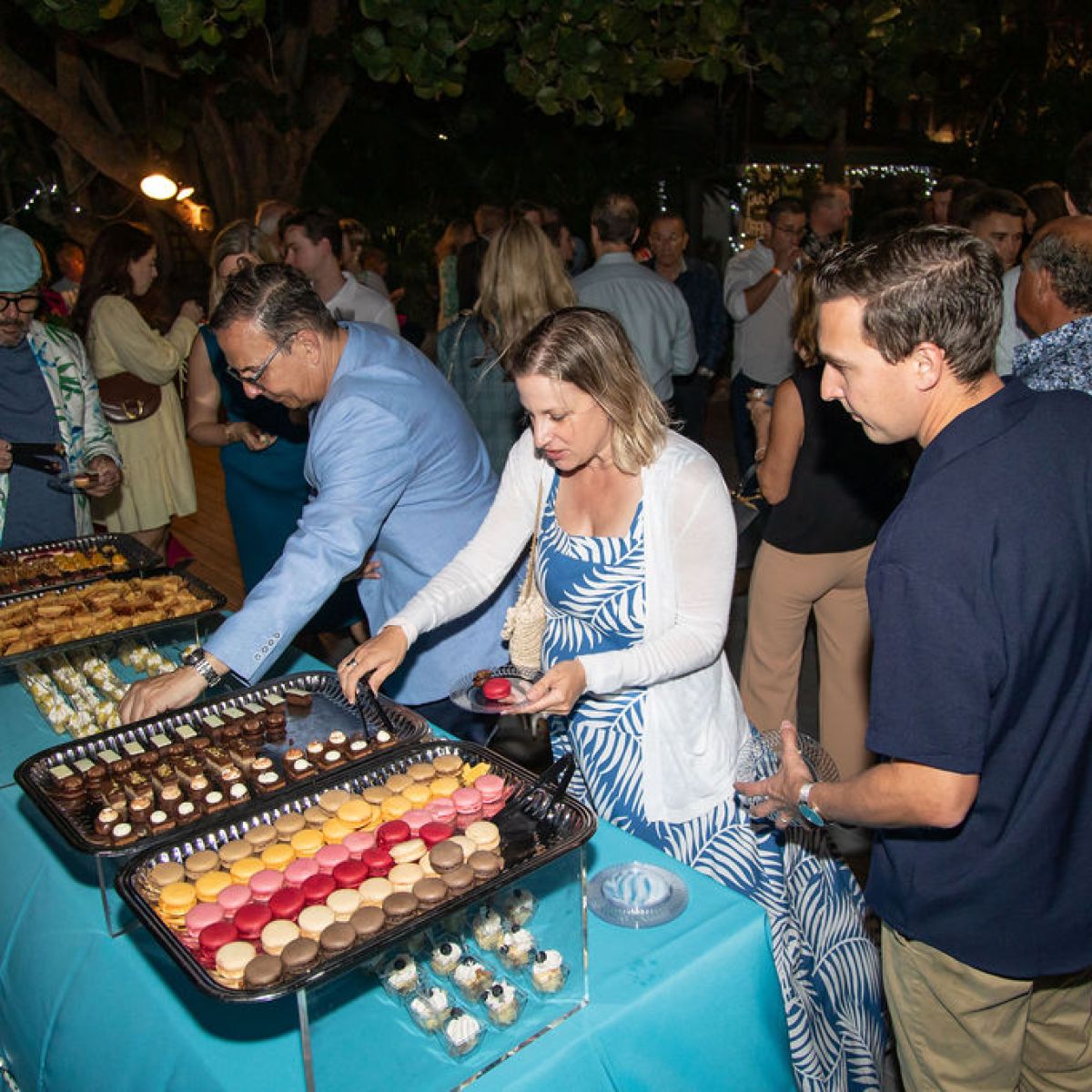 People at a party buffet table with various desserts, including macarons, outdoors at night.