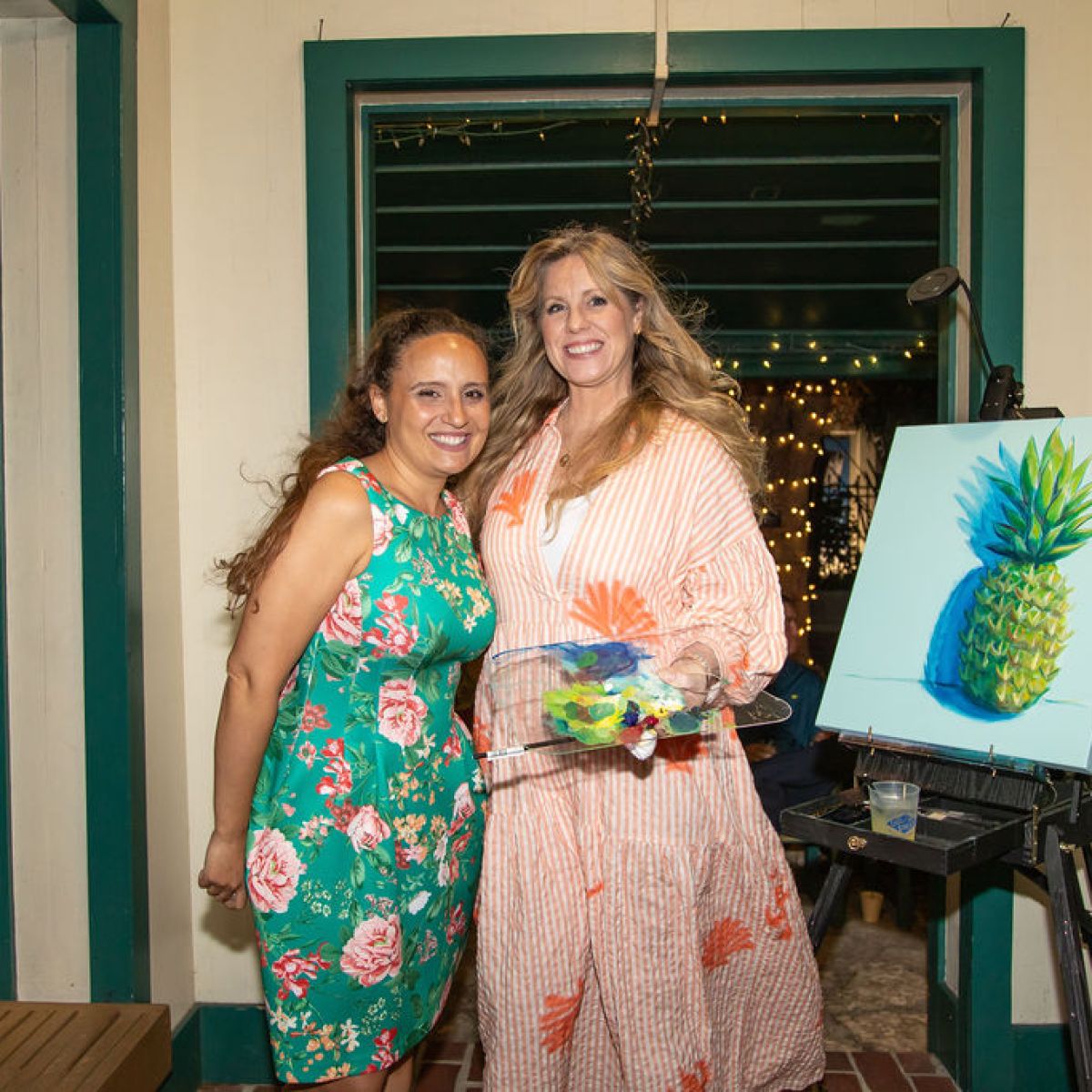 Two women smiling, one holding a paint palette, near a pineapple painting on an easel.