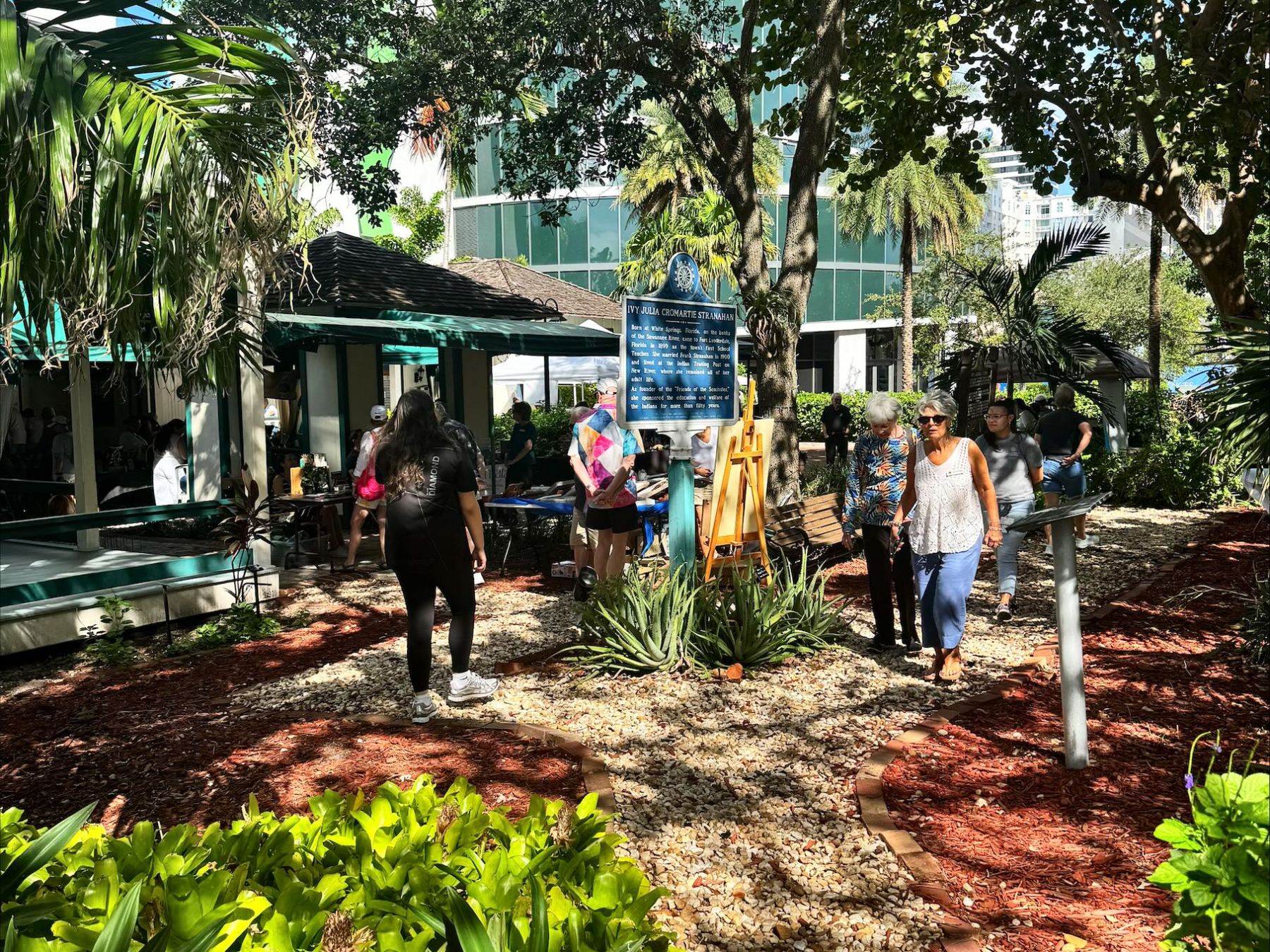 People walking on a garden path surrounded by trees and plants under a clear blue sky.