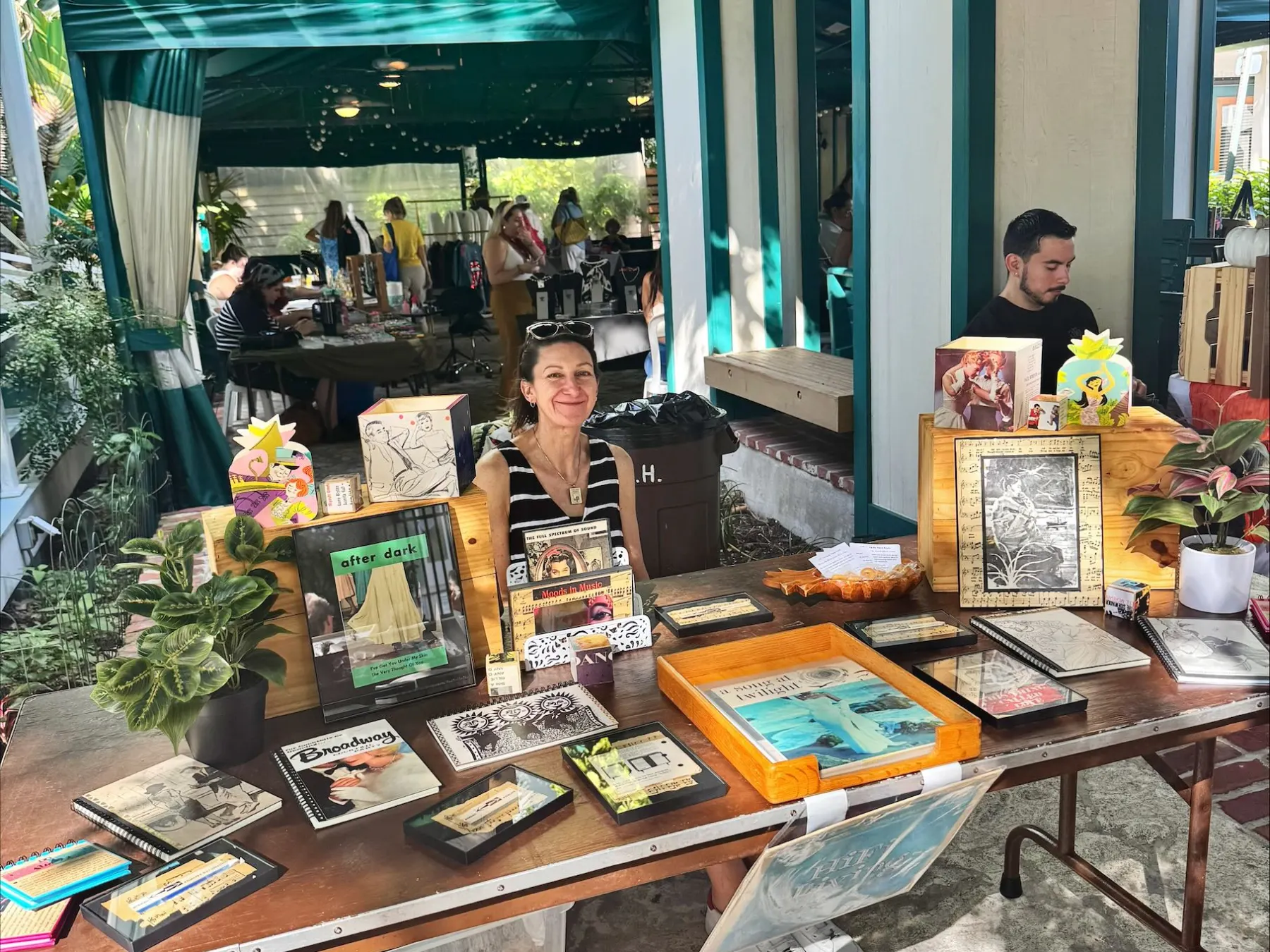 Outdoor market stall with art and crafts display, two people behind the table.