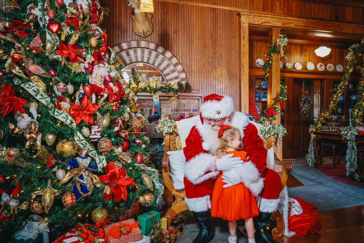 Santa hugging a child in a red dress by a decorated Christmas tree indoors.