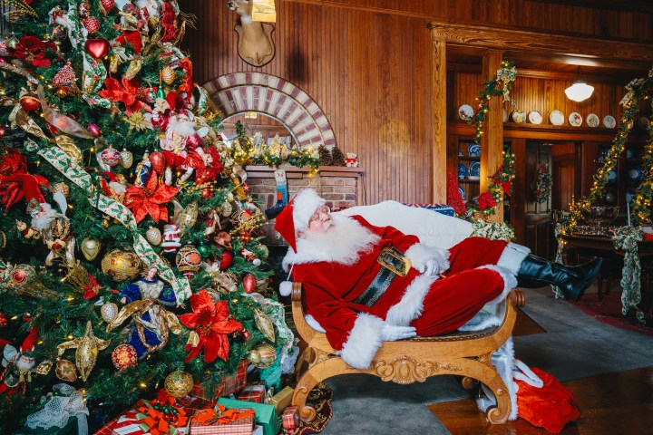 Santa Claus sleeps on a sofa beside a decorated Christmas tree in a festive room.