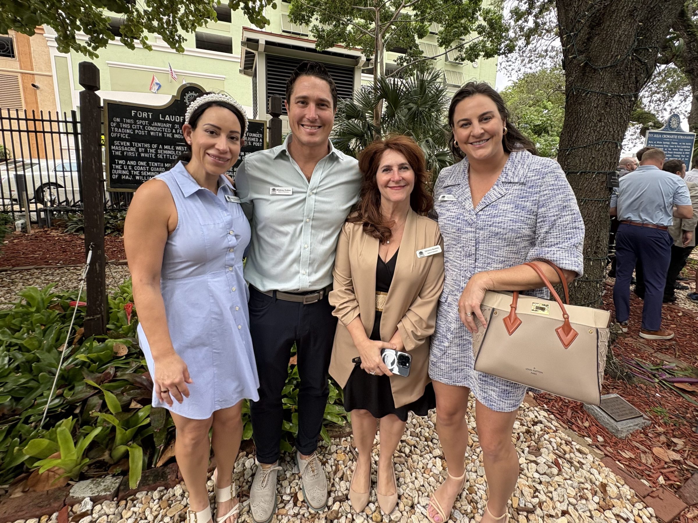 Four people smiling outdoors near a historical marker, with trees and a crowd in the background.