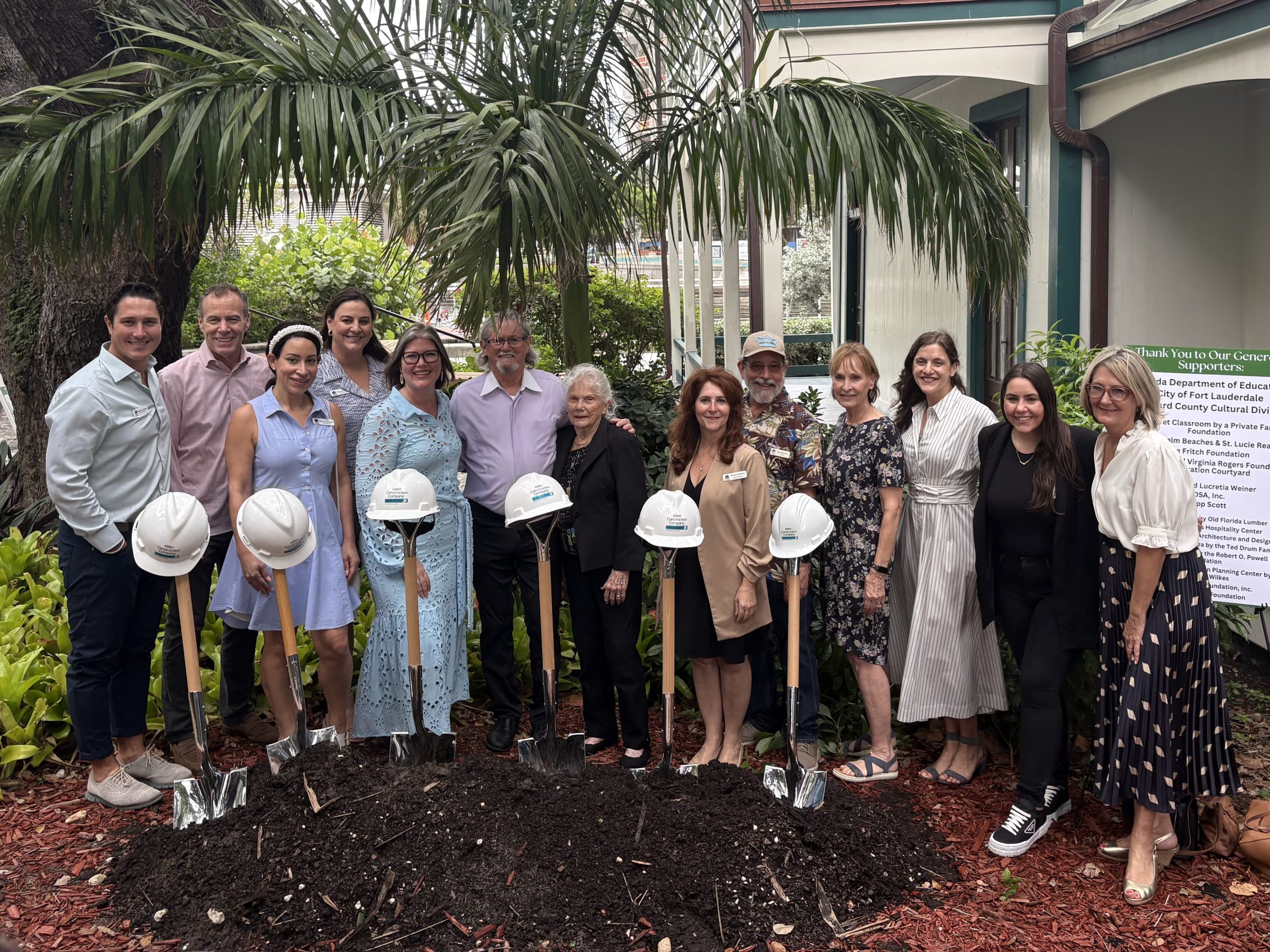 Group of people posing with shovels and hard hats in a garden outside a house.