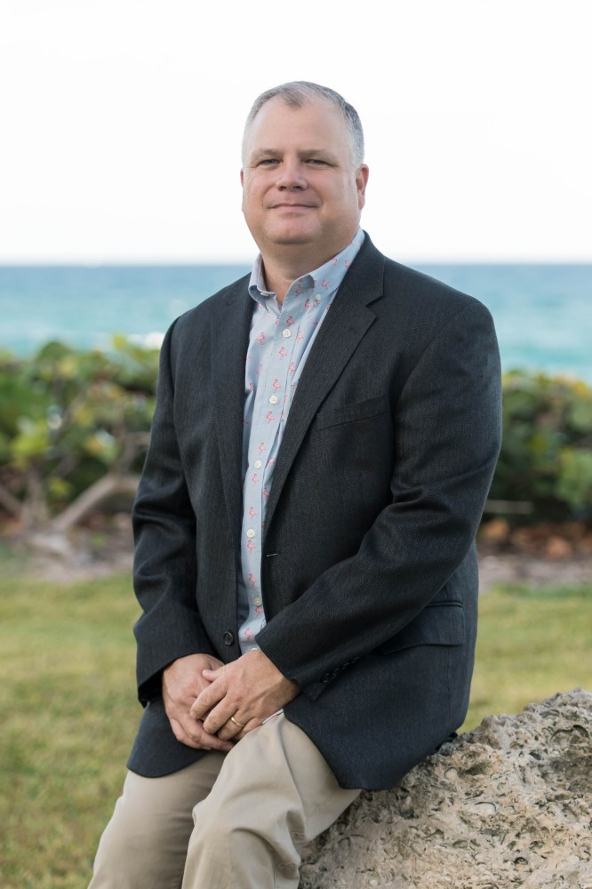 Man in blazer sits on rock by ocean.